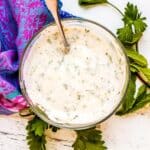 Bowl of Vegan Yogurt Sauce with a spoon it in next to sprigs of fresh parsley and mint.