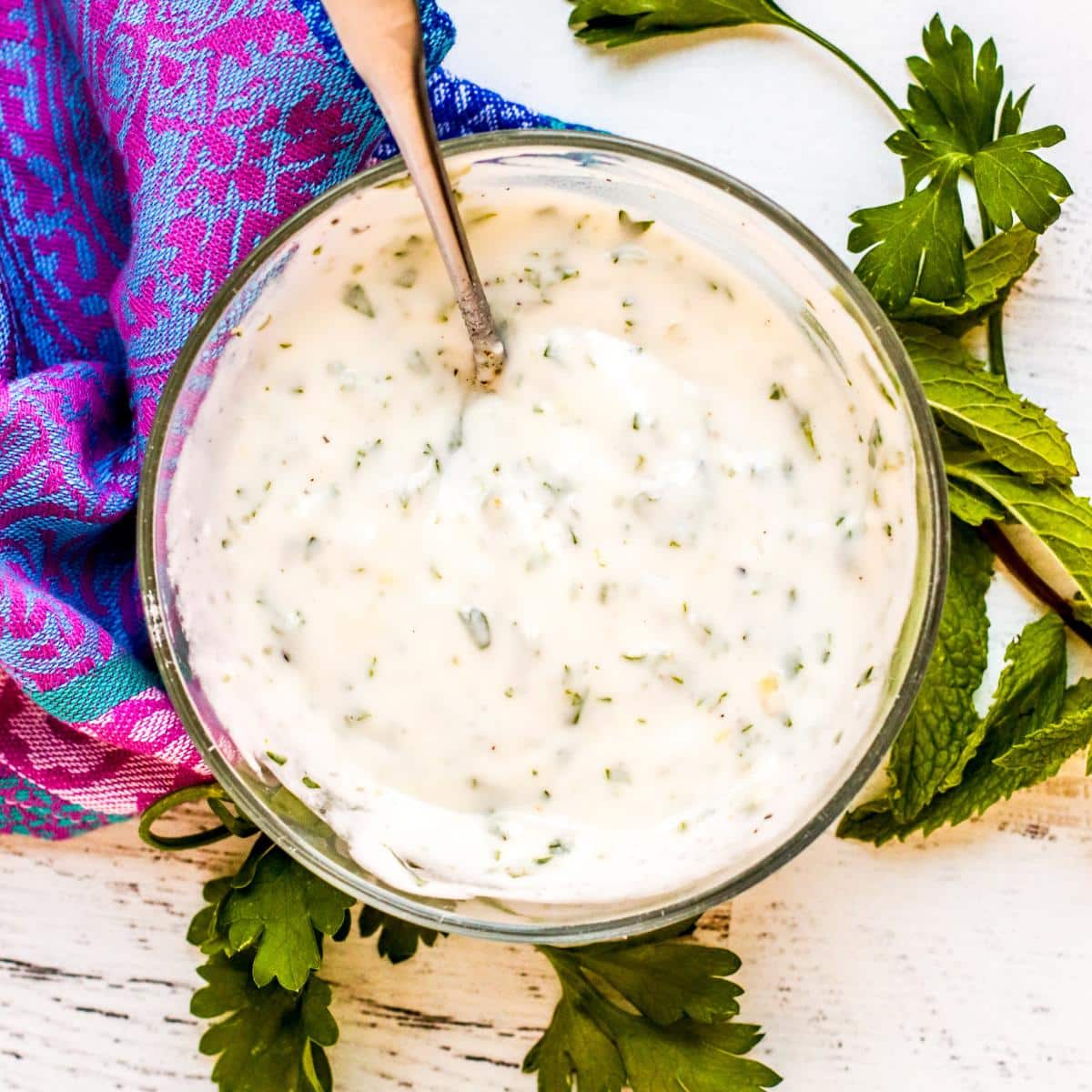 Bowl of Vegan Yogurt Sauce with a spoon it in next to sprigs of fresh parsley and mint.