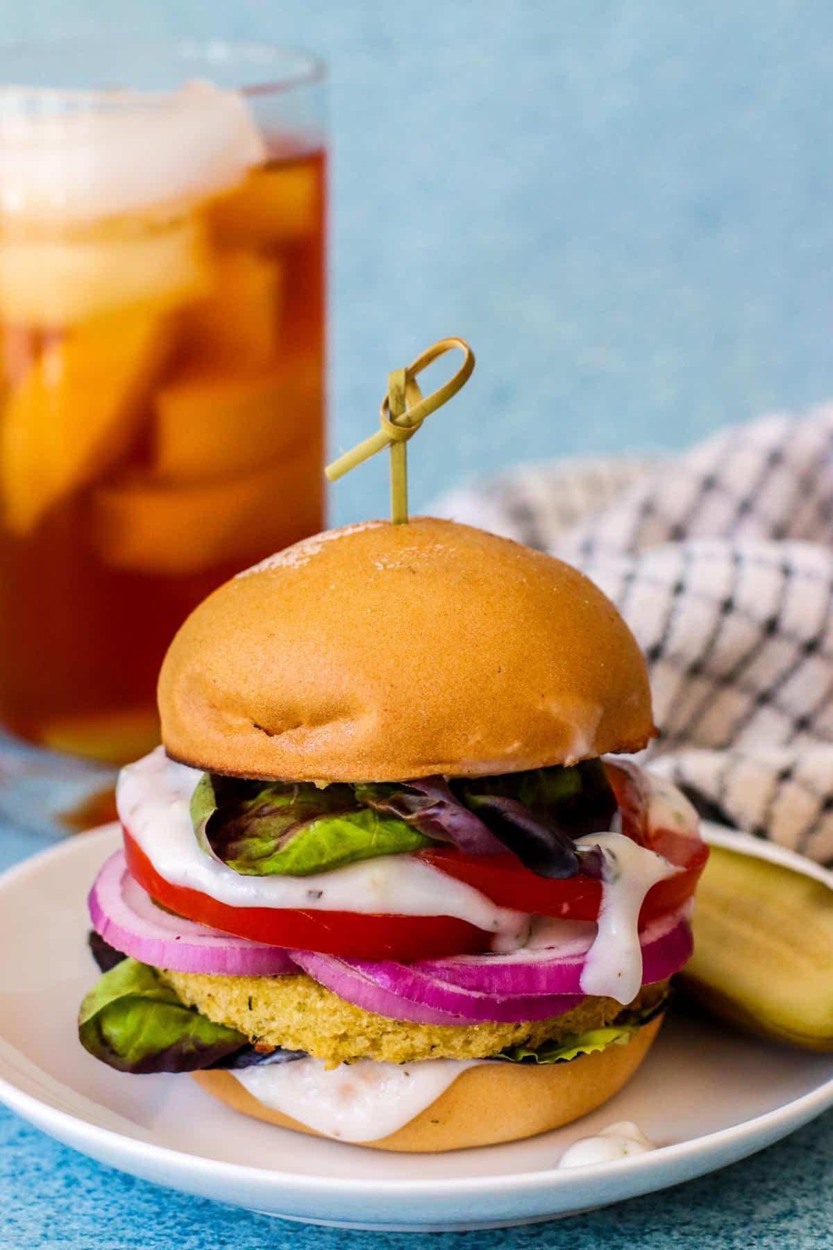 Veggie burger with lettuce, tomatoes, red onions, and yogurt sauce with a glass of iced tea in the background.