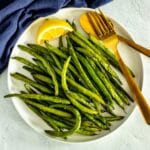 Platter of green beans with a wedge of lemon and gold serving utensils.
