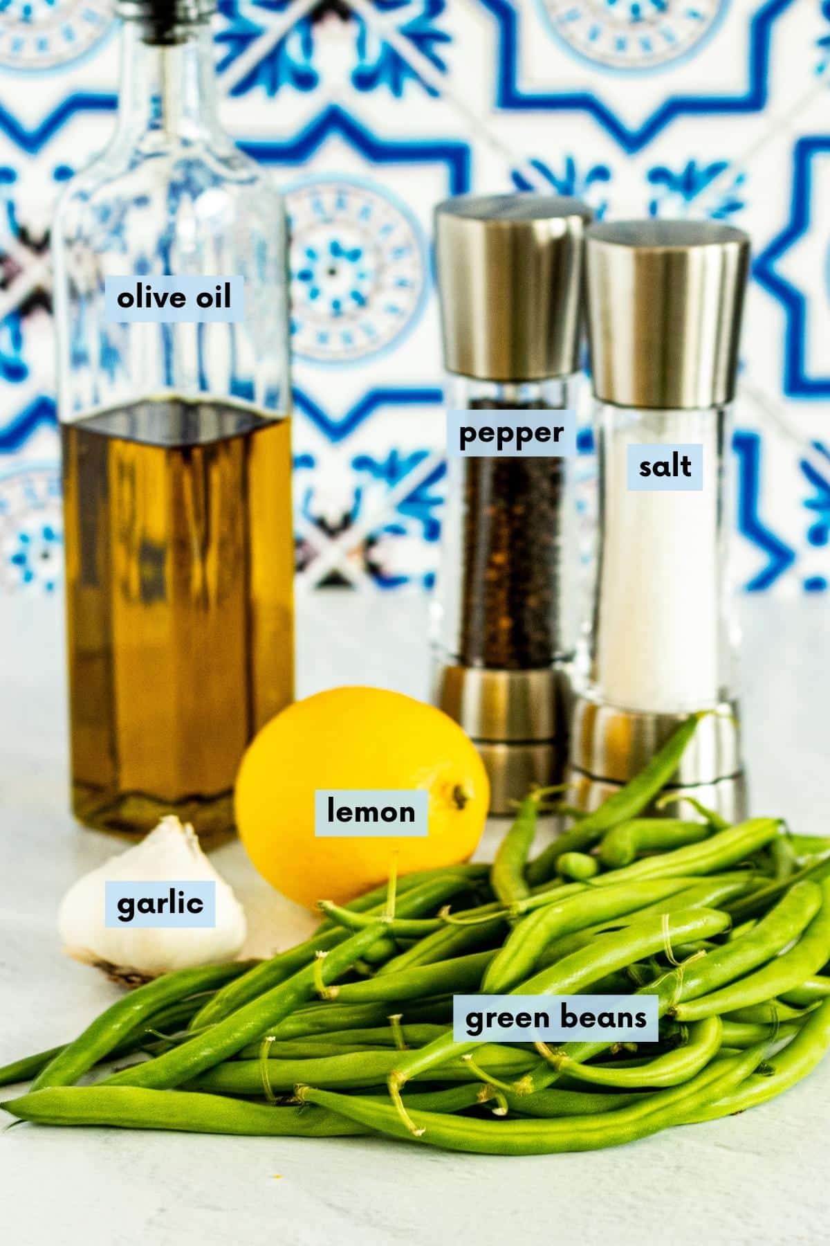 Fresh green beans, garlic, lemon, bottle of olive oil, and salt and pepper grinders on a kitchen counter.