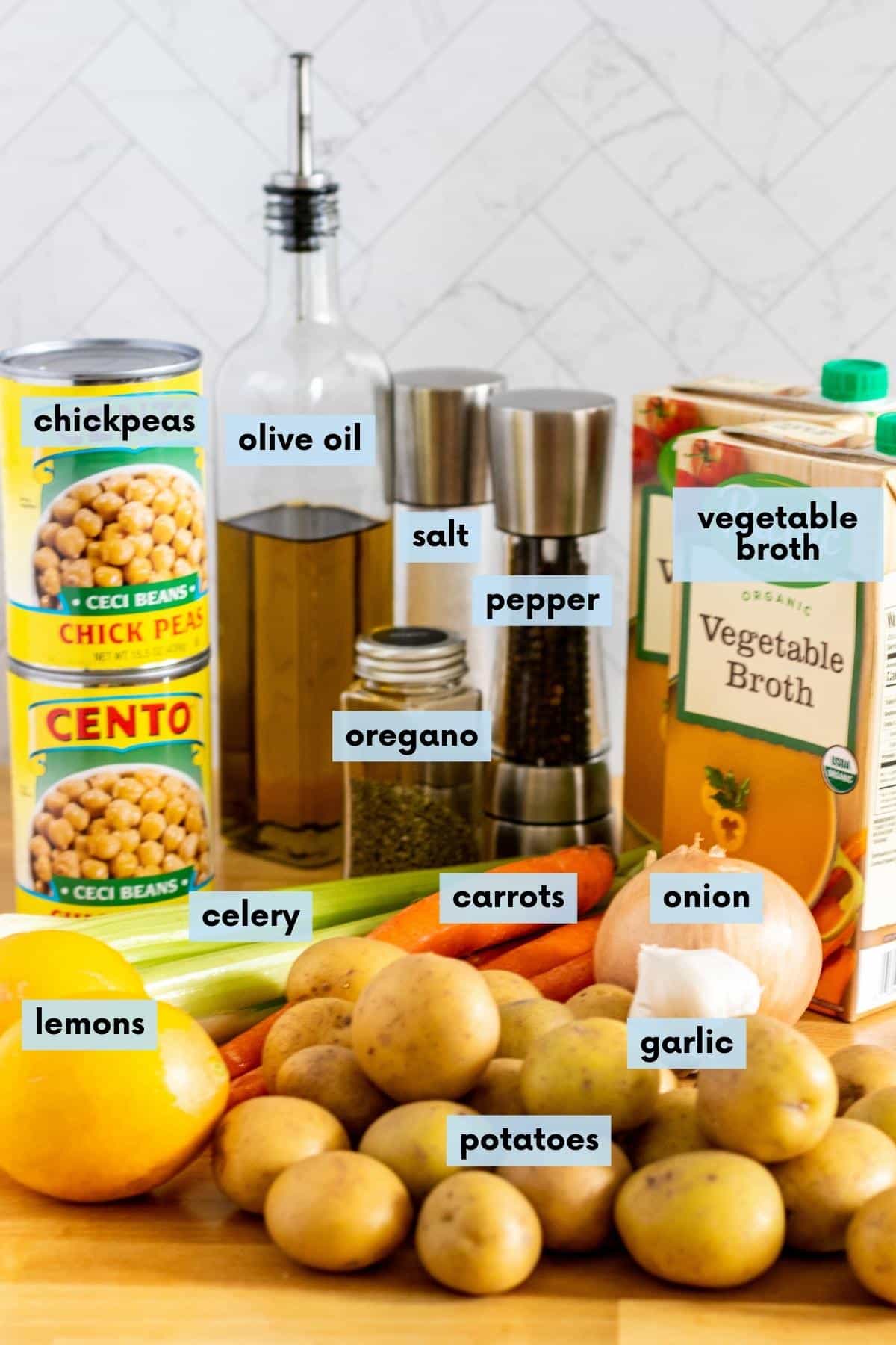 Potatoes, lemons, celery, carrots, onion, garlic, cans of chickpeas, bottle of olive oil, jar of dried oregano, salt and pepper grinders, and boxes of vegetable broth on a kitchen counter.