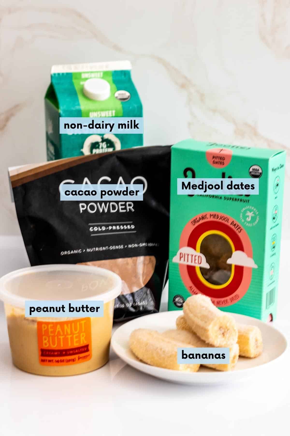 Container of peanut butter, plate with frozen banana halves, bag of cacao powder, carton of non-dairy milk, and box of Medjool dates on a kitchen counter.