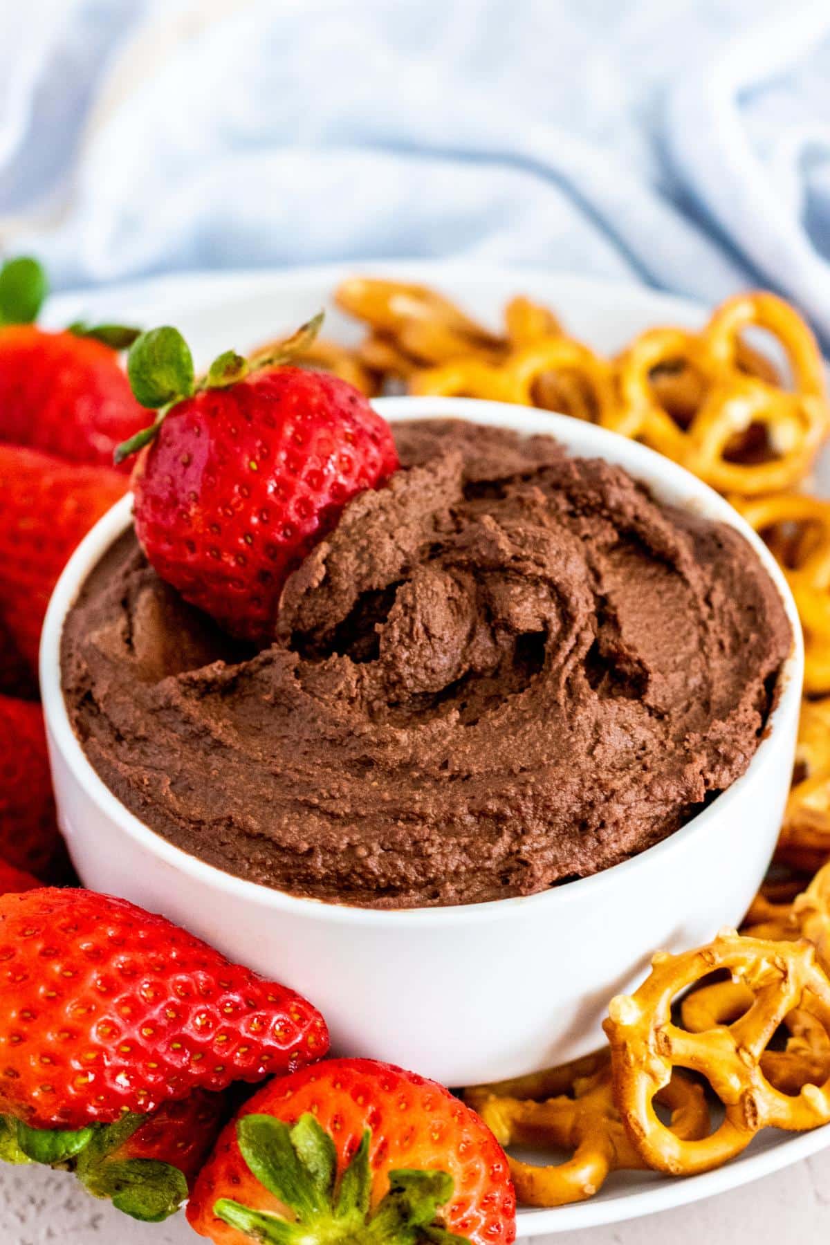 Bowl of dark chocolate hummus with a strawberry being dipped it in surrounded by more strawberries and mini pretzels.