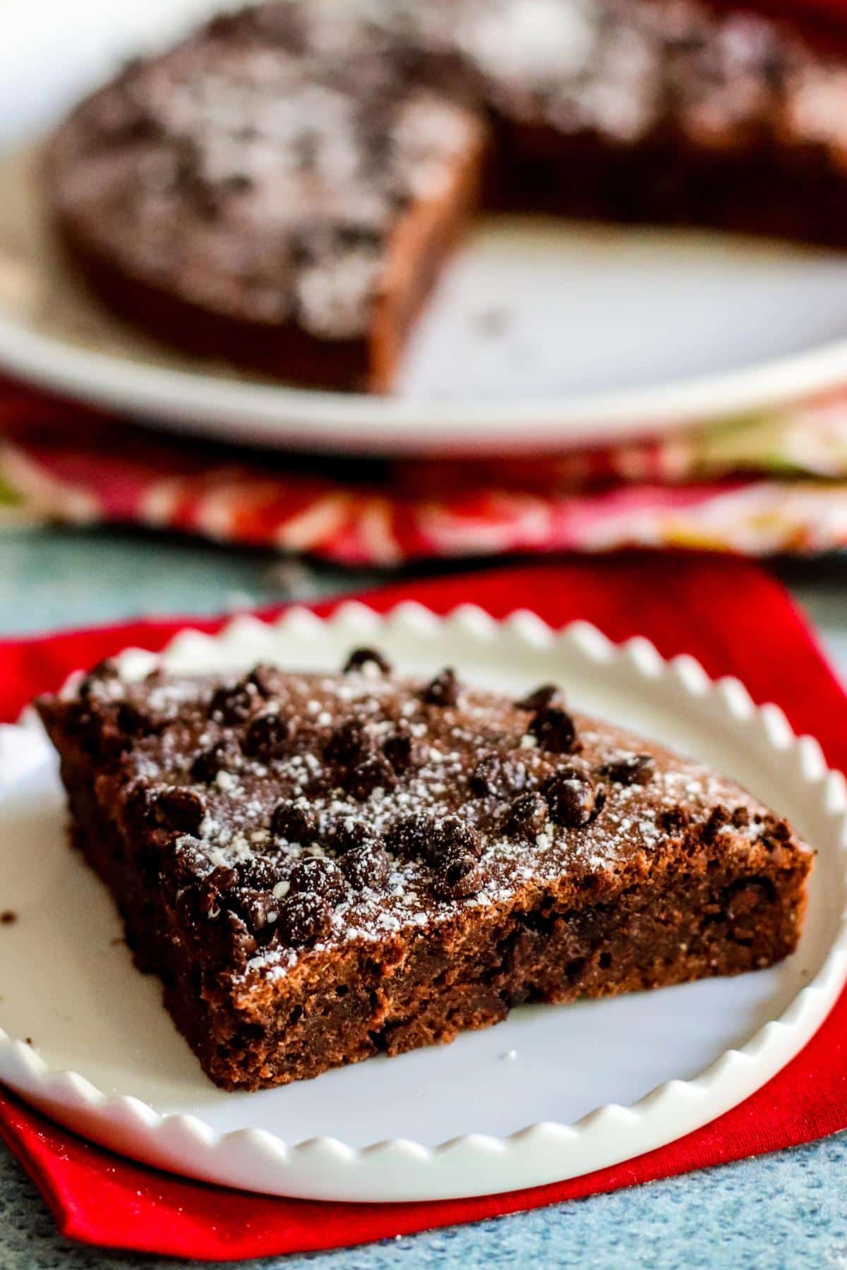 Brownie on a plate topped with mini chocolate chips and powdered sugar with platter in the background.