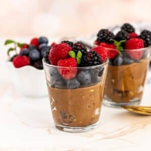 Glasses of chocolate mousse topped with fresh berries and a bowl of berries in the background.