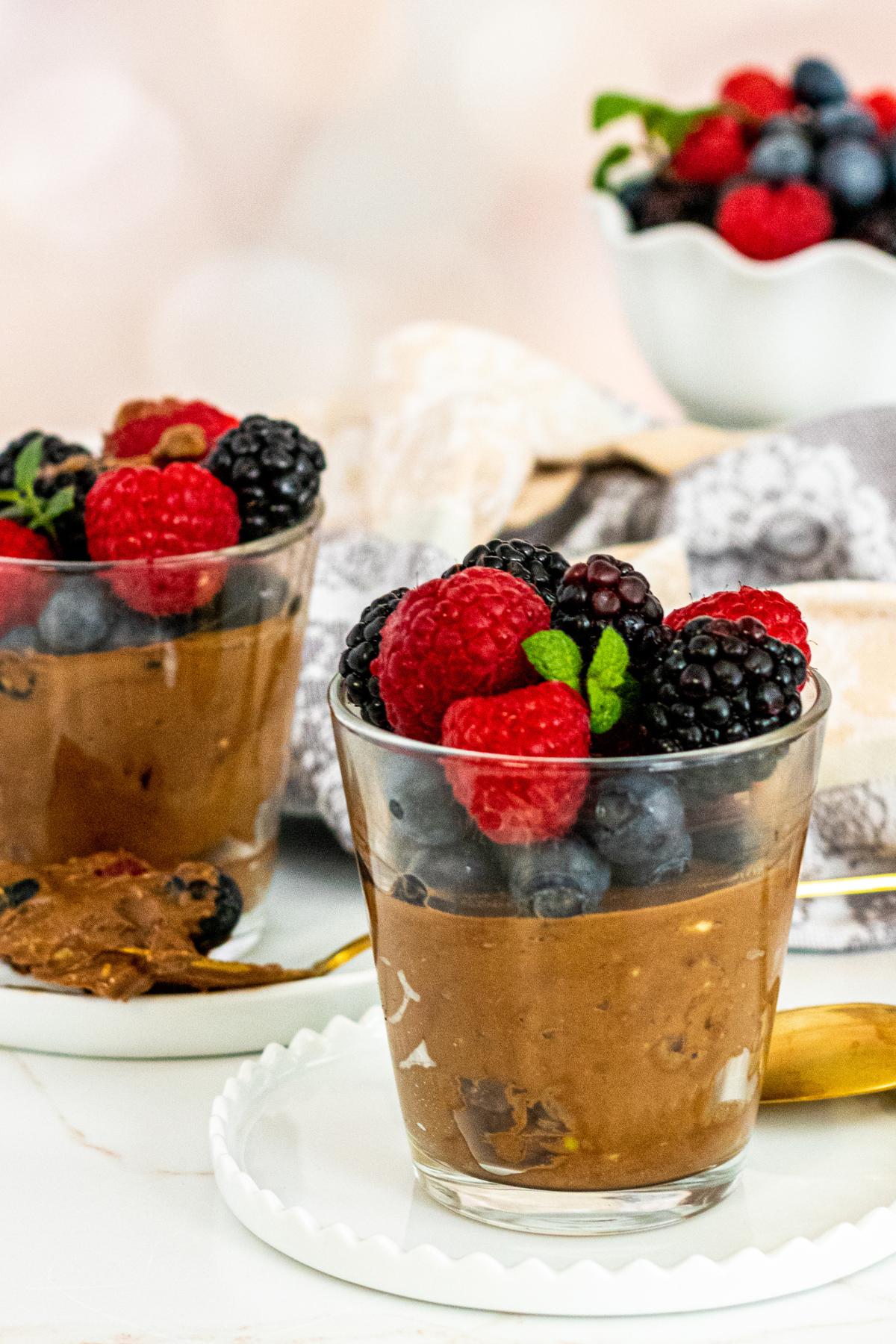 Glasses of mousse topped with berries and a bowl of berries in the background.