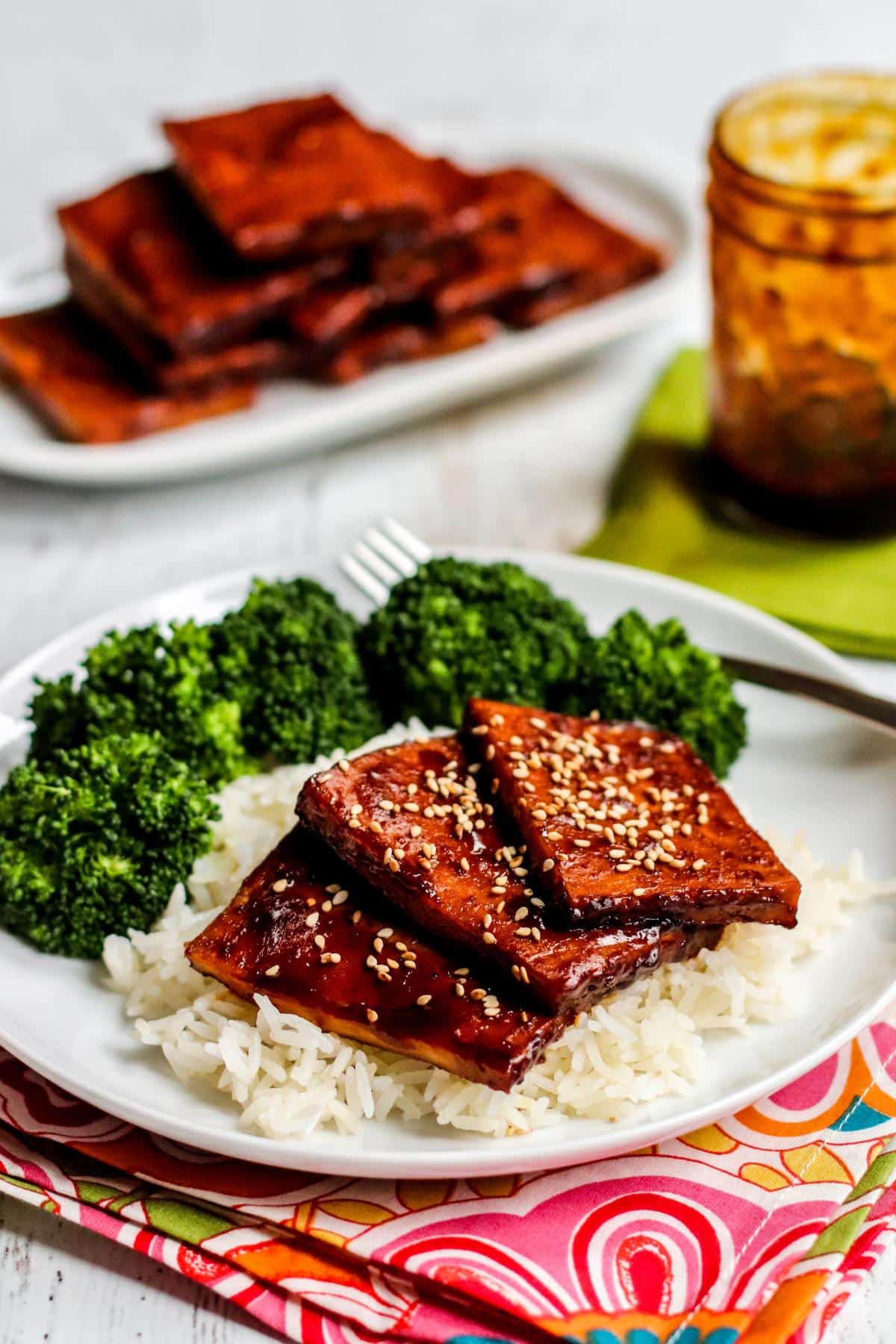 Plate with slices of tofu on rice with steamed broccoli with a platter of baked teriyaki tofu and a jar of sauce in the background.
