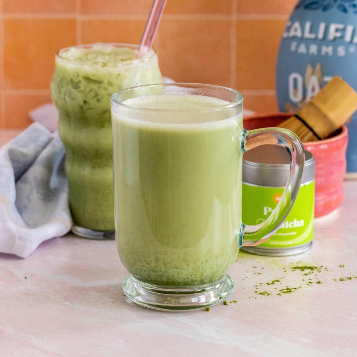 Glasses of hot and iced oat milk matcha lattes with container of matcha, a bowl and bamboo whisk, and bottle of oat milk in the background,.