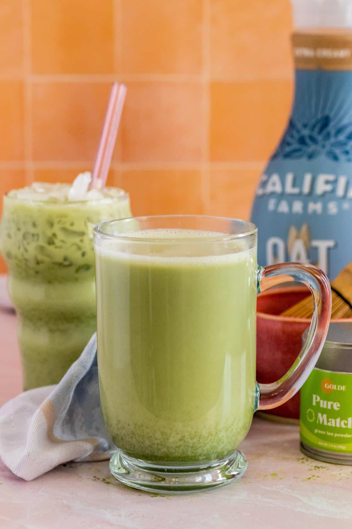 Glasses of hot and iced matcha lattes with tin of matcha powder, a bowl and whisk, and a bottle of oat milk in the background.