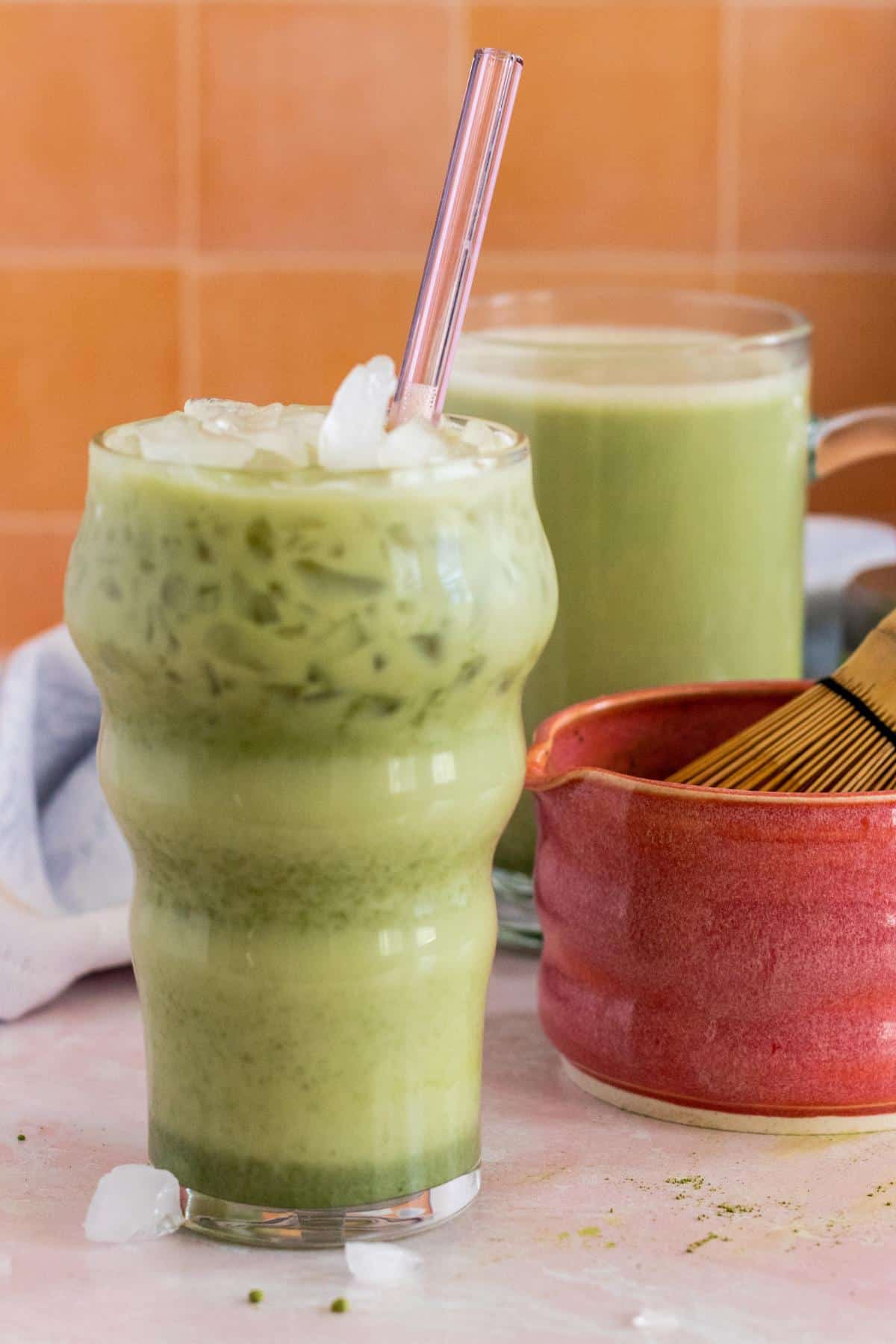 A glass of iced matcha latte with a pink glass straw and a coral ceramic bowl of mug of hot matcha latte in the background.