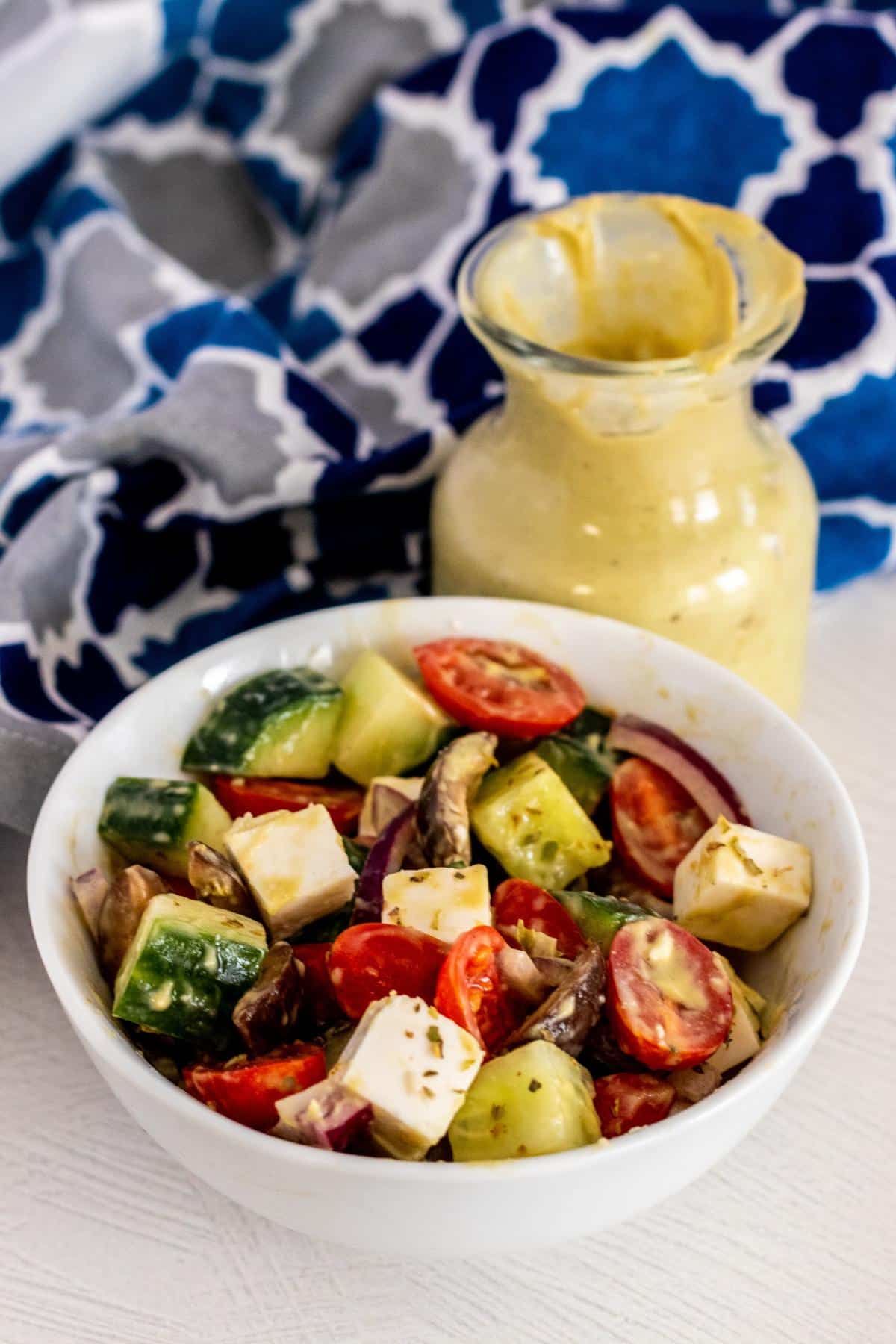 Bowl of vegan Greek salad made with vegan feta cubes, cucumbers, tomatoes, red onions, and black olives with bottle of avocado dressing next to it and blue patterned napkin.
