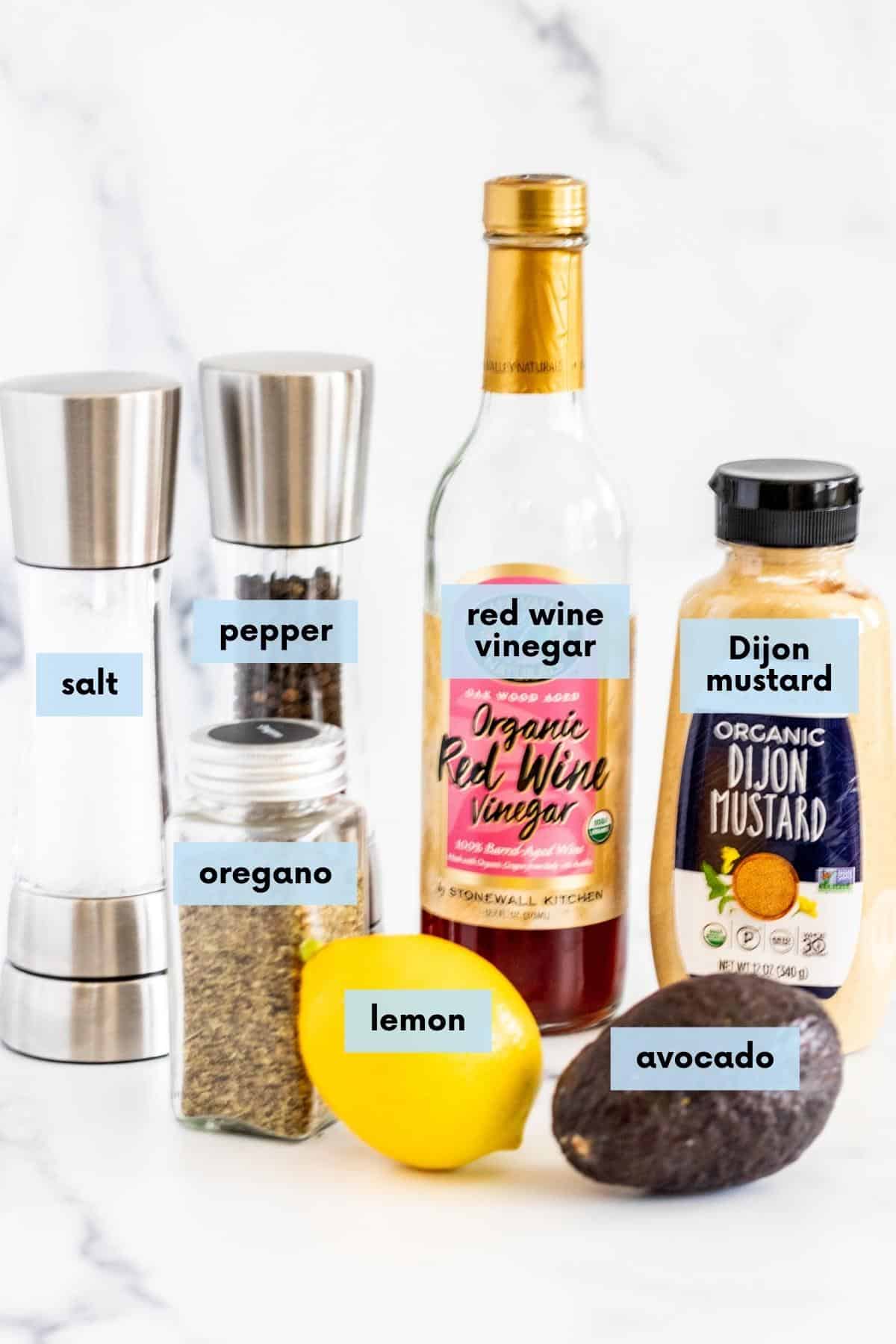 Salt and pepper shakers, jar of dried oregano, bottle of red wine vinegar, bottle of Dijon mustard, a lemon, and an avocado.