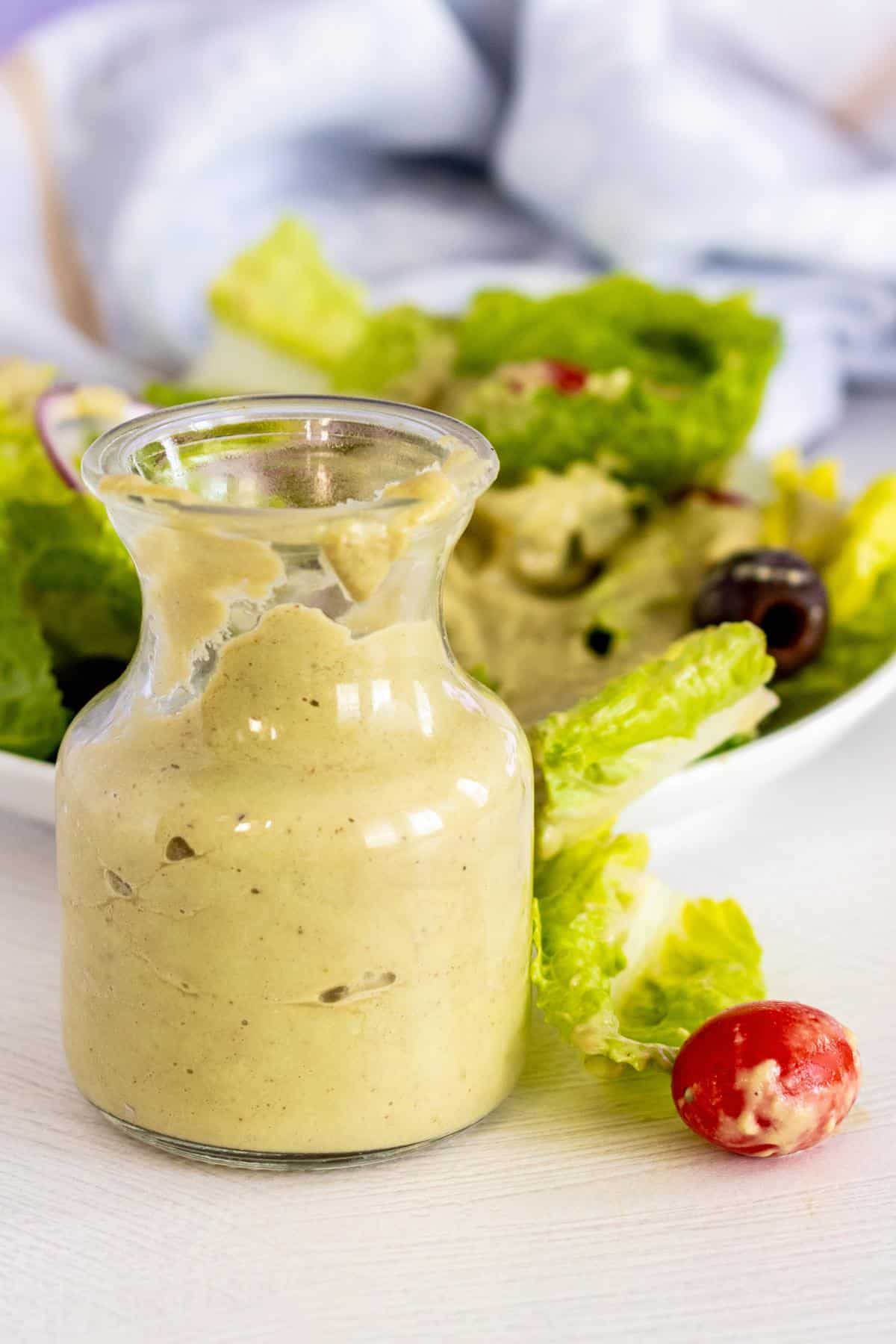 Vegan Avocado Dressing in a bottle next to a salad drizzled with dressing and a cherry tomato that fell off the plate.