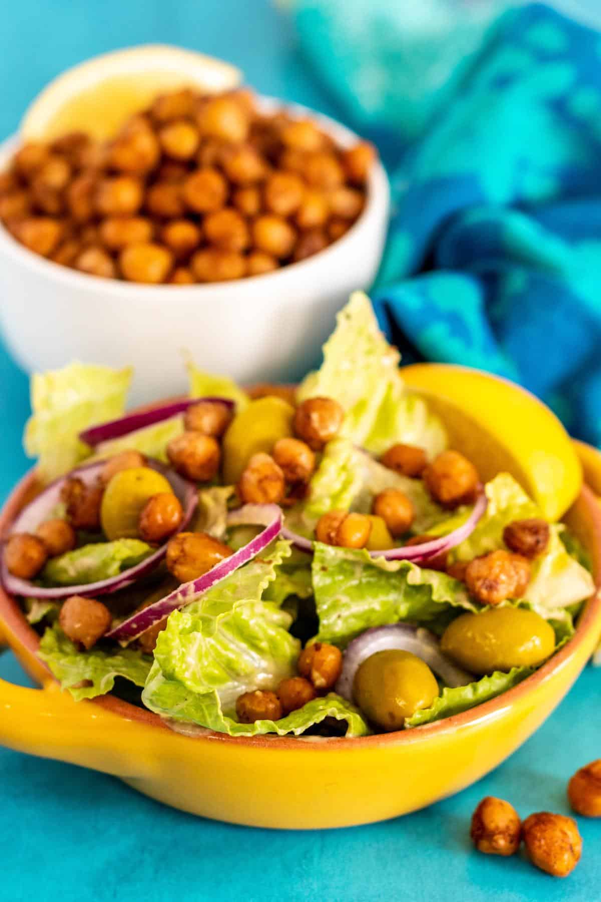Salads with lettuce, green olives, red onion, and chickpeas with bowl of pan fried chickpeas in the background.