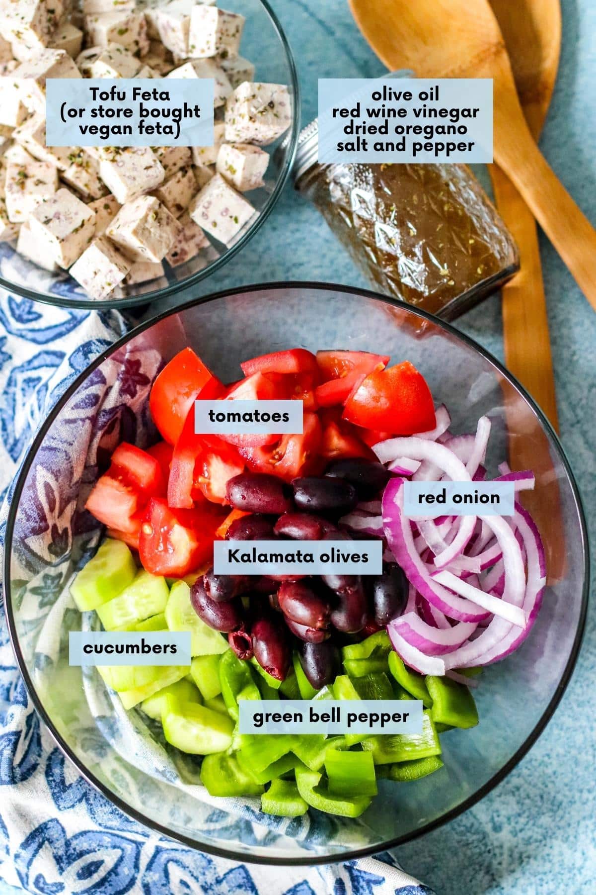Large salad bowl with tomatoes, cucumbers, green bell peppers, red onions, and Kalamata olives, small bowl of vegan feta cheese, small jar of dressing, and salad tongs.