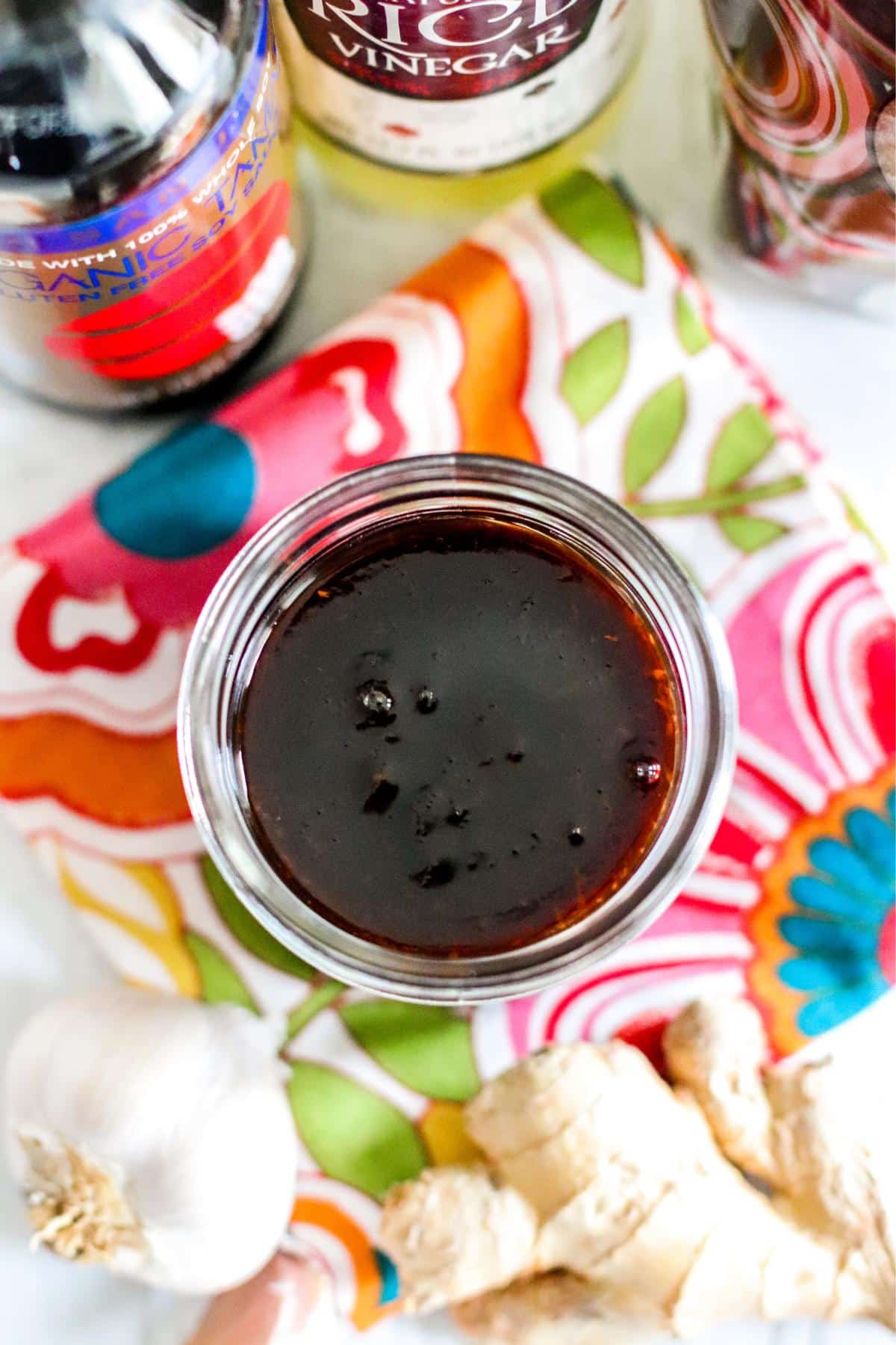 Sauce in a jar surrounded by bottles of tamari and rice vinegar, a bag of coconut sugar, and fresh ginger and garlic.