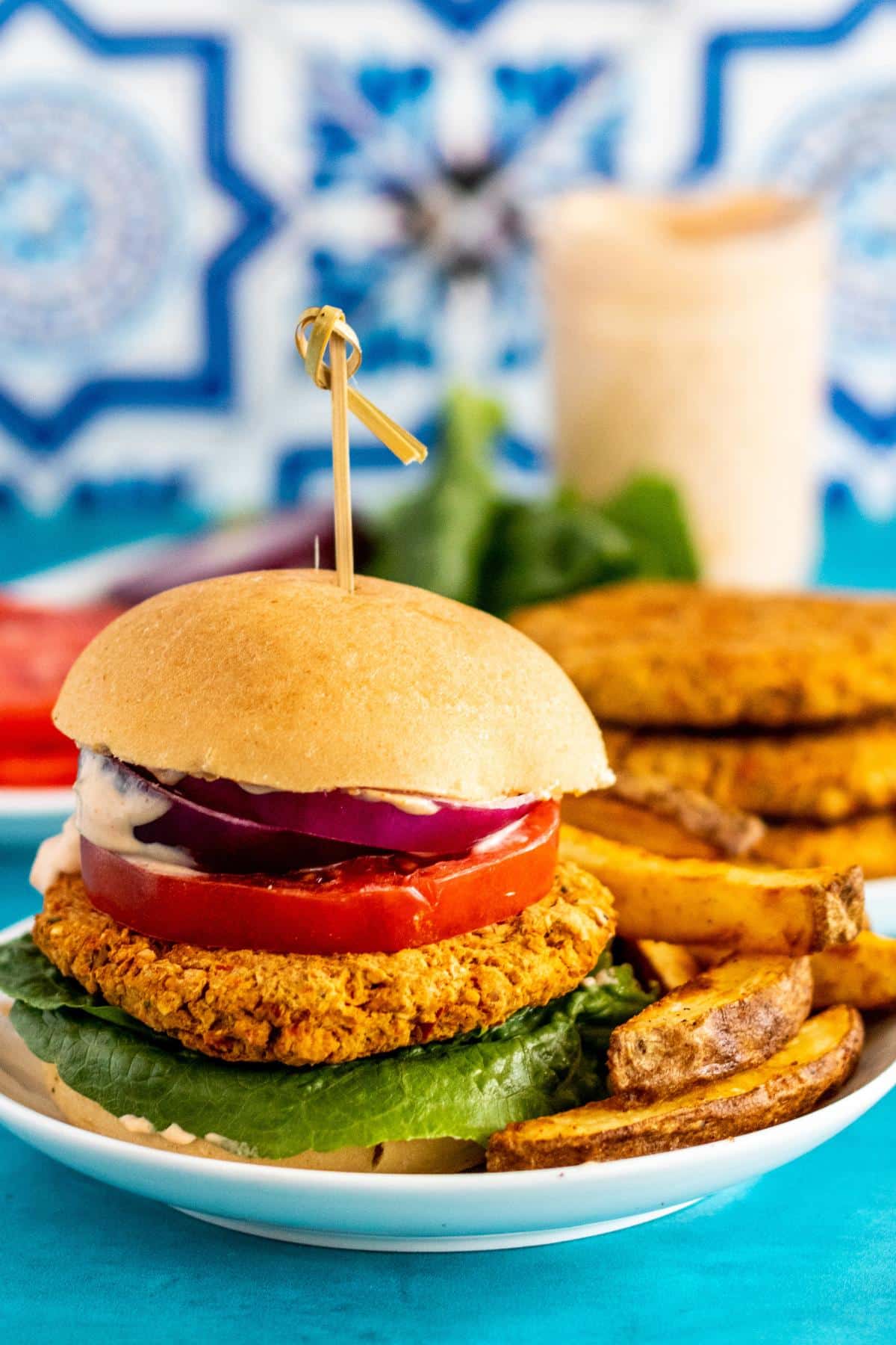 A chickpea burger on a bun with lettuce, tomato, and red onion with a side of fries and a plate of patties and jar of sauce in the background.