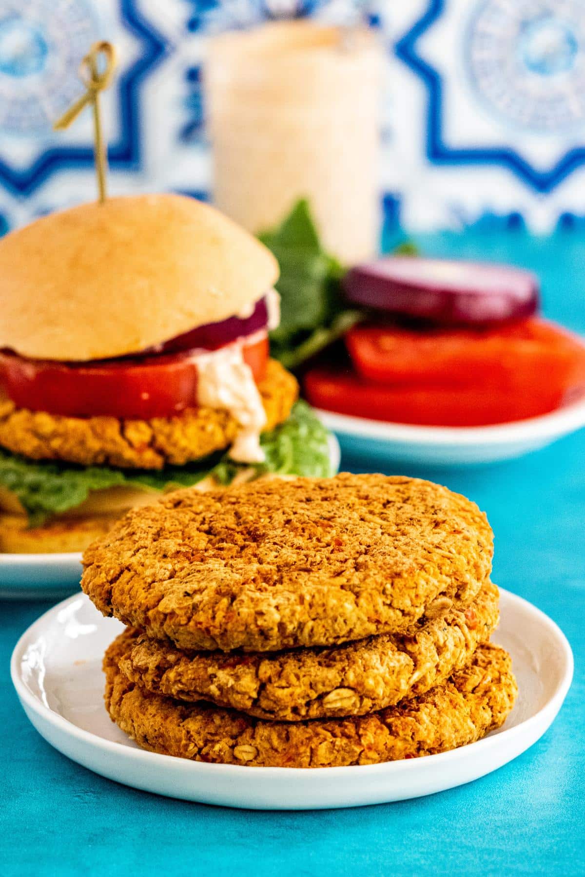 3 chickpea burger patties on a plate with a chickpea burger, plate of lettuce, tomato and onion slices, and a jar of sauce in the background.