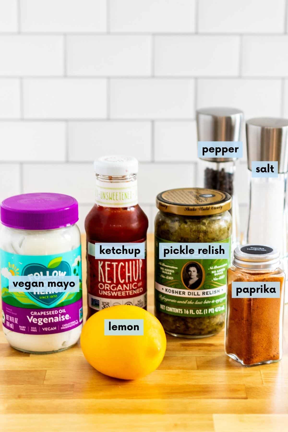 Jar of vegan mayo, bottle of ketchup, jar of pickle relish, jar of paprika, salt and pepper grinders, and a lemon on a kitchen counter.