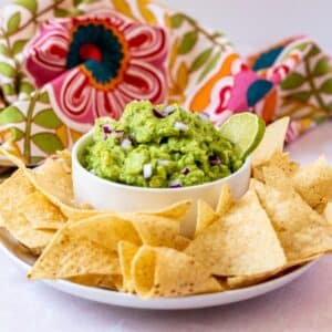 Bowl of guacamole on a platter surrounded by tortilla chips with a floral cloth napkin in the background.