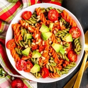 Bowl of red and green pasta topped with cherry tomatoes, avocado, and pine nuts.