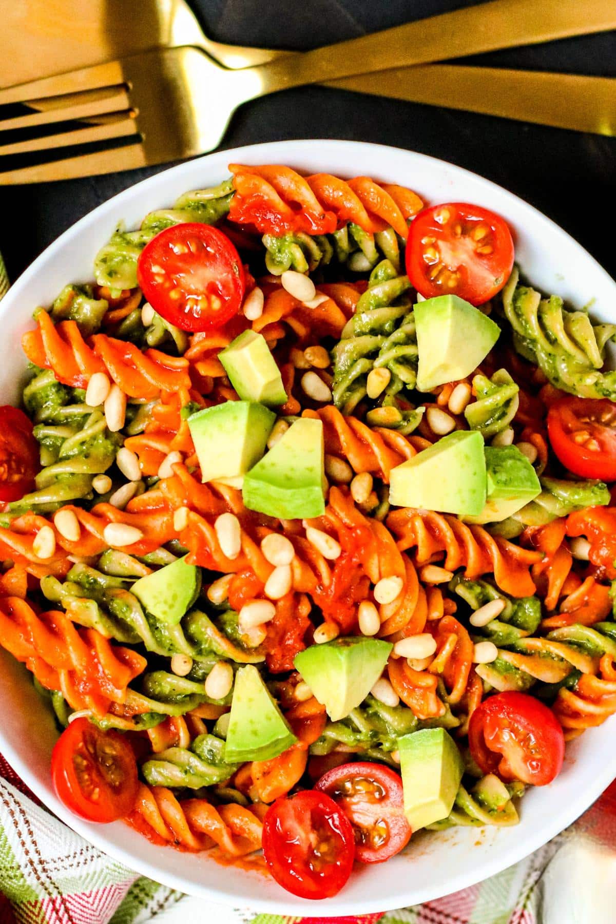 Bowl of rotini pasta tossed with red and green sauces topped with cherry tomatoes, avocado, and pine nuts with gold serving utensils.