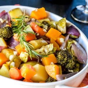 Roasted vegetables in a white casserole dish garnished with a sprig of fresh rosemary.