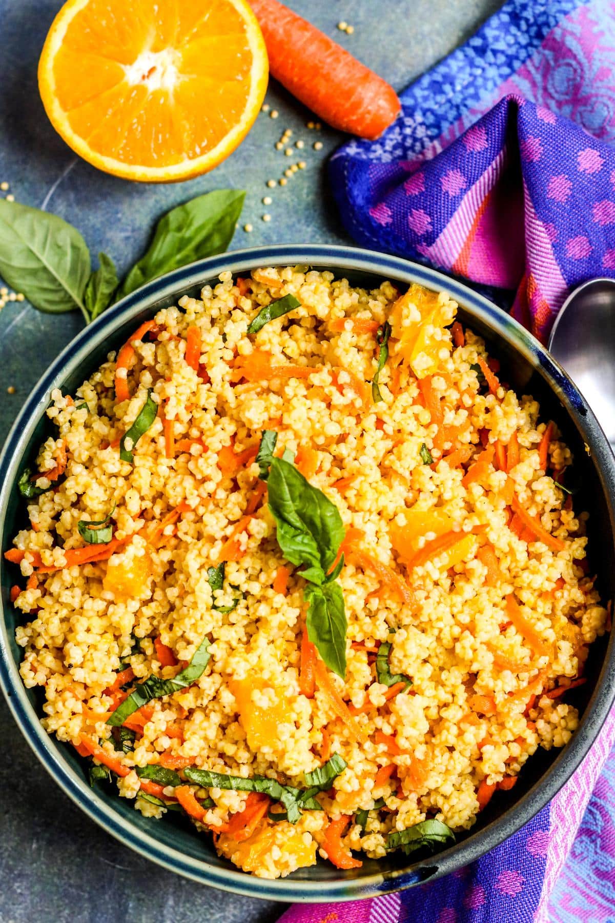 Bowl of millet salad next to half an orange, a carrot, and fresh basil leaves.