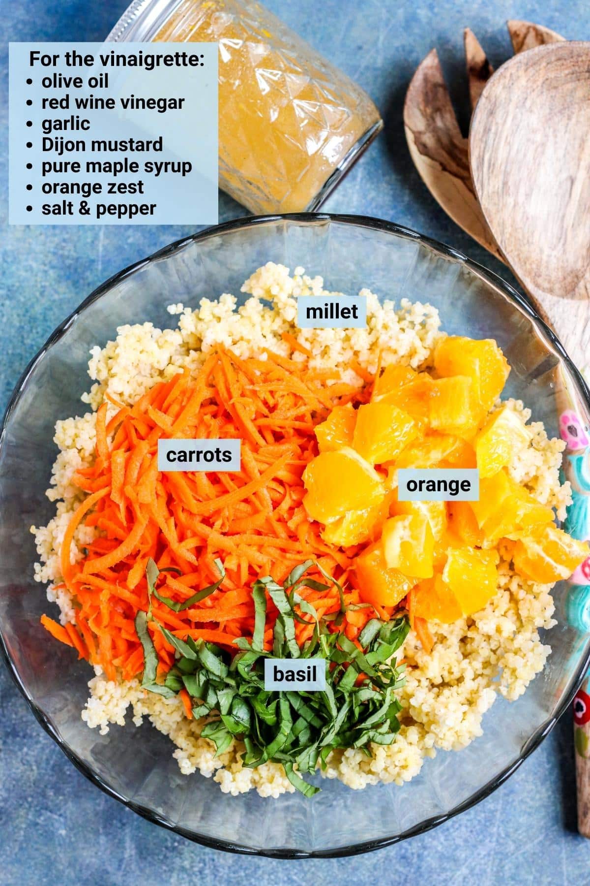Bowl with cooked millet, shredded carrots, orange segments, and fresh basil ribbons with a jar of dressing and salad serving utensils next to it.