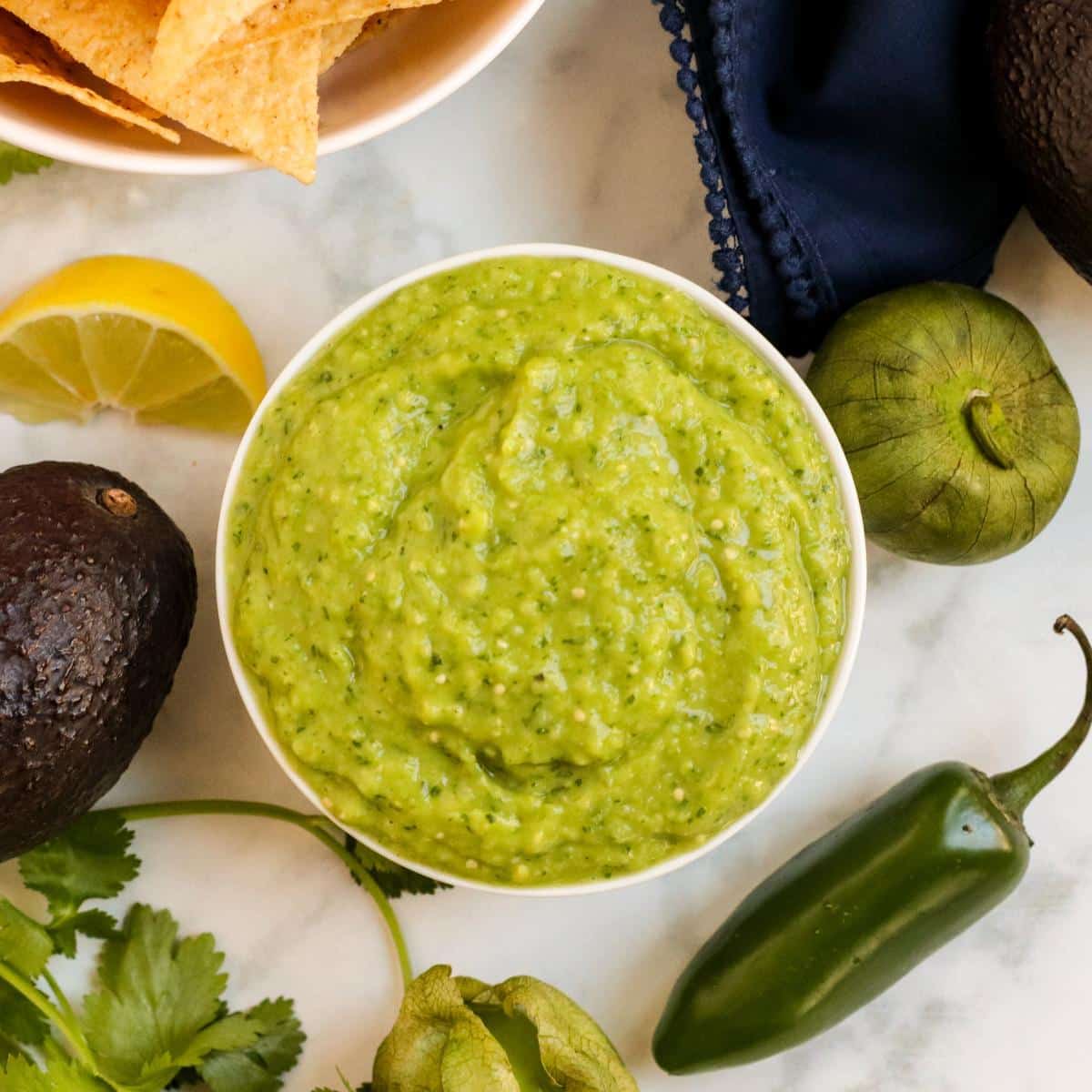 Bowl of green salsa, an avocado, jalapeno pepper, cilantro, tomatillo, and sprig of cilantro.