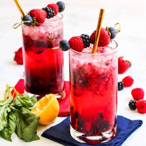Red cocktails in tall glasses garnished with blueberries, raspberries, blackberries, and strawberries.