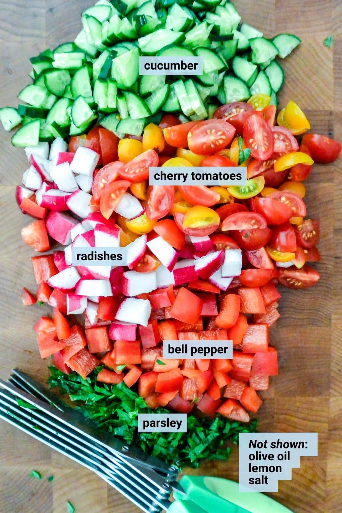 Diced cucumbers, tomatoes, radishes, bell peppers, and chopped parsley with an herb scissors on a cutting board.