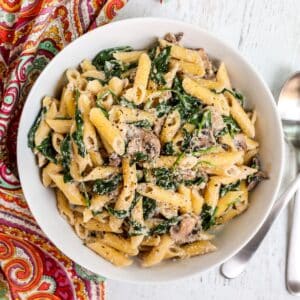 Bowl of penne pasta with spinach and mushrooms in a creamy white sauce.