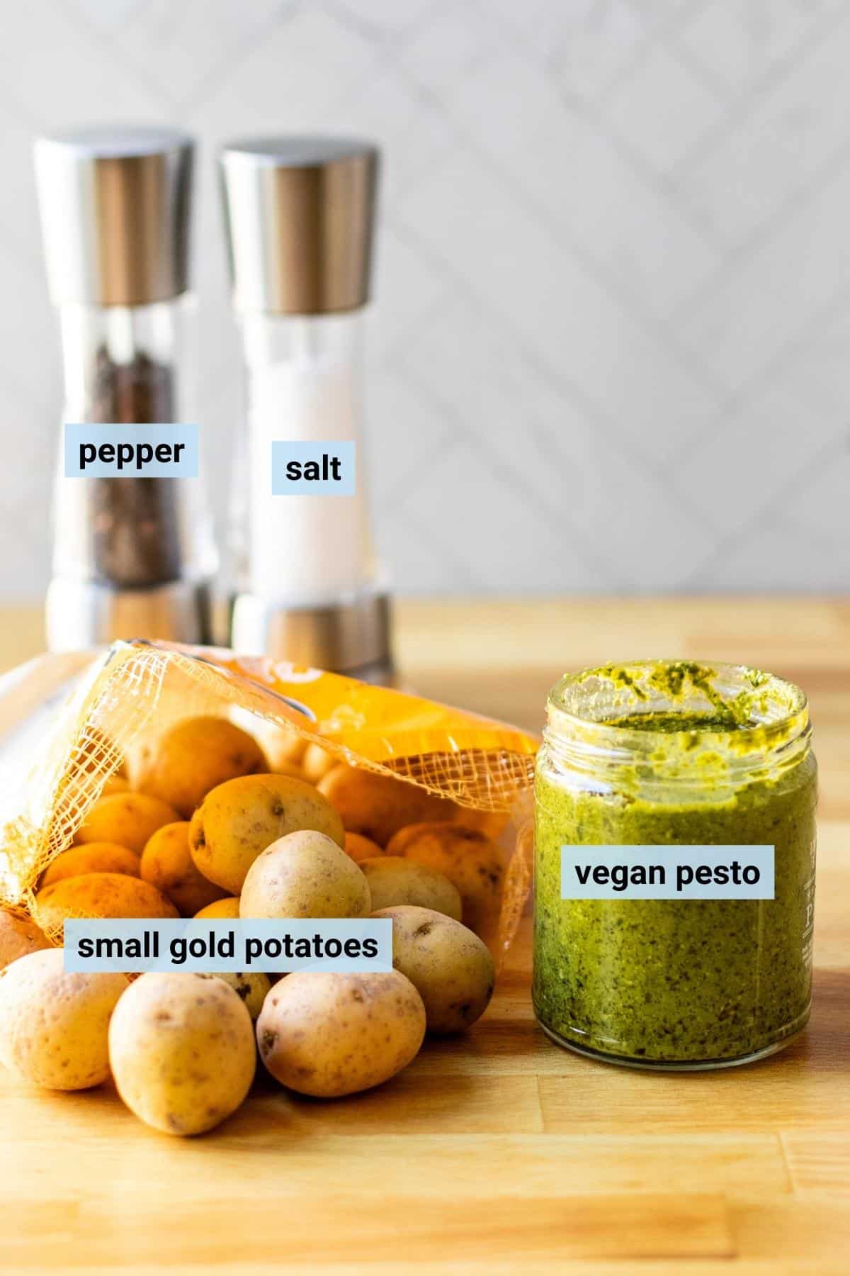 Bag of small gold potatoes, jar of vegan pesto, and salt and pepper grinders on a cutting board.