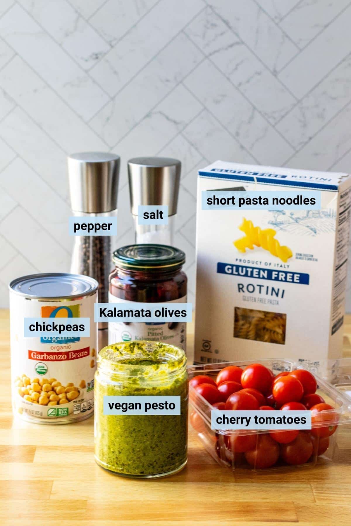 Can of chickpeas, jar of vegan pesto sauce, basket of cherry tomatoes, jar of Kalamata olives, box of rotini pasta, and salt and pepper grinders.