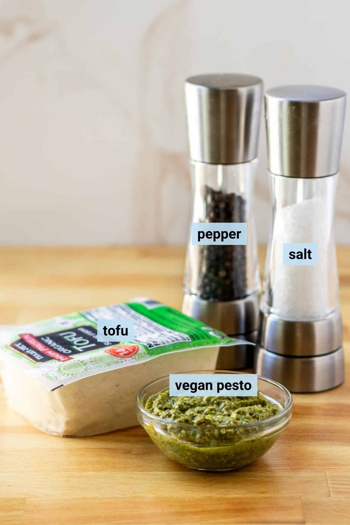 Bowl of vegan pesto sauce, block of super-firm tofu, and salt and pepper grinders on a cutting board.