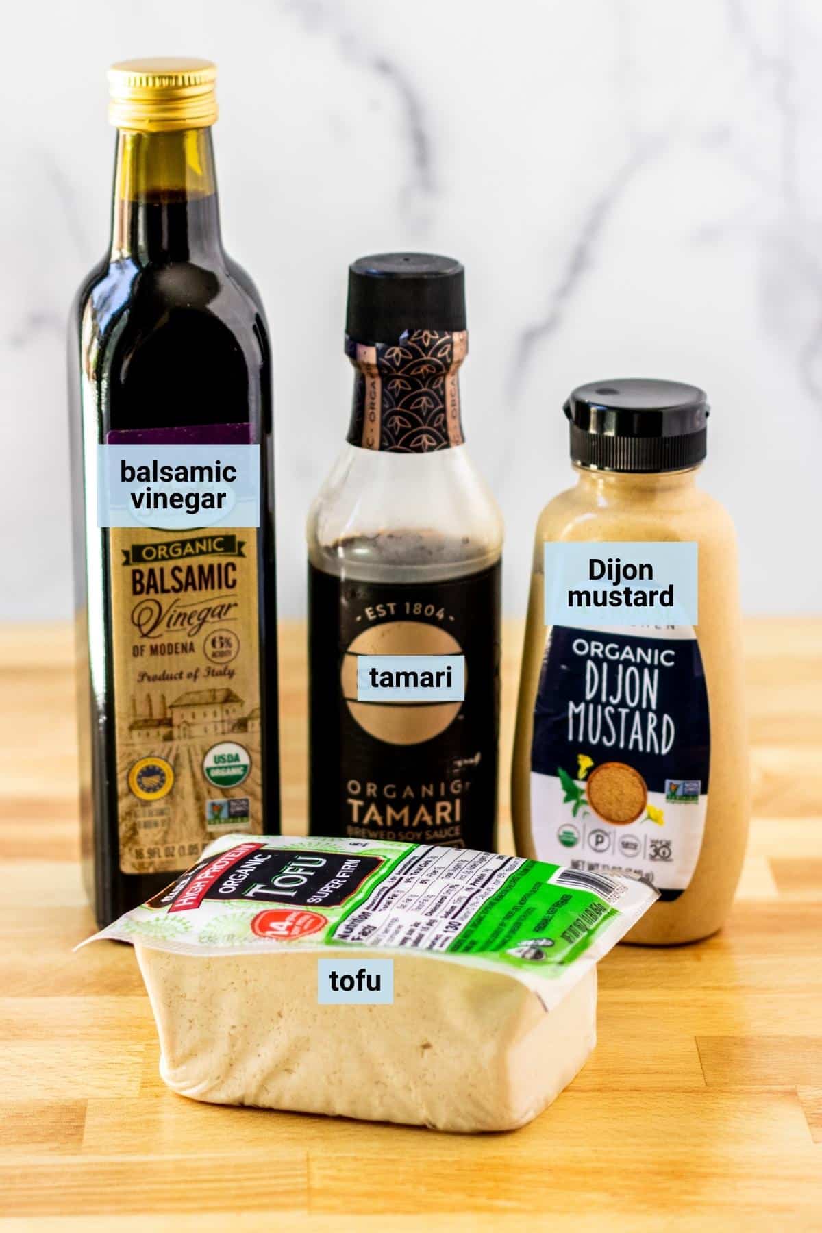 Block of tofu and bottles of balsamic vinegar, tamari, and Dijon mustard on a cutting board.