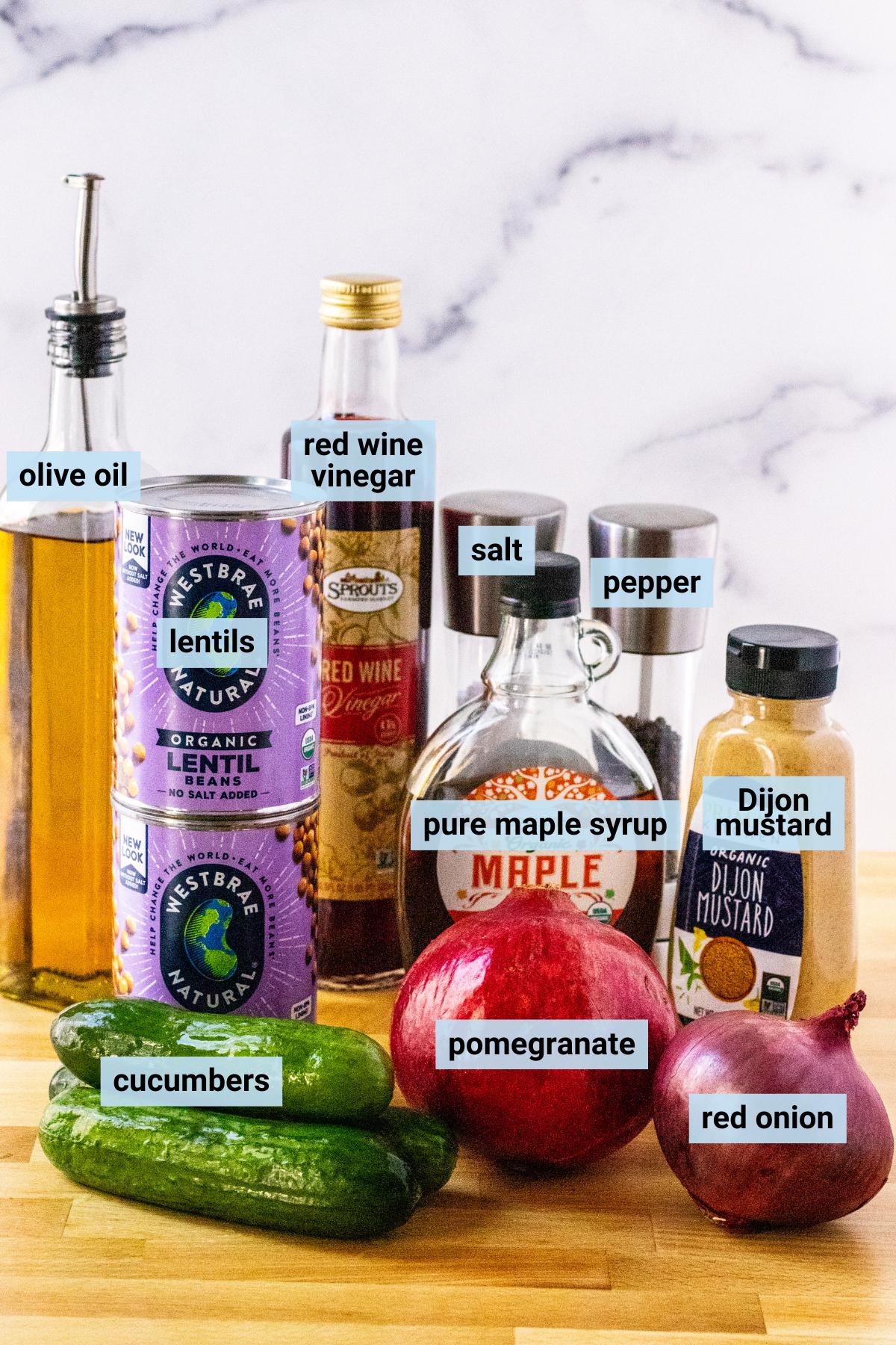 Cucumbers, pomegranate, red onion, cans of lentils, bottle of olive oil, bottle of red wine vinegar, bottle or pure maple syrup, jar of Dijon mustard, and salt and pepper grinders.