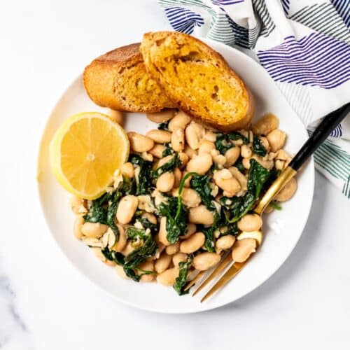 Spinach and white beans on a plate with toasted baguette and a lemon wedge.