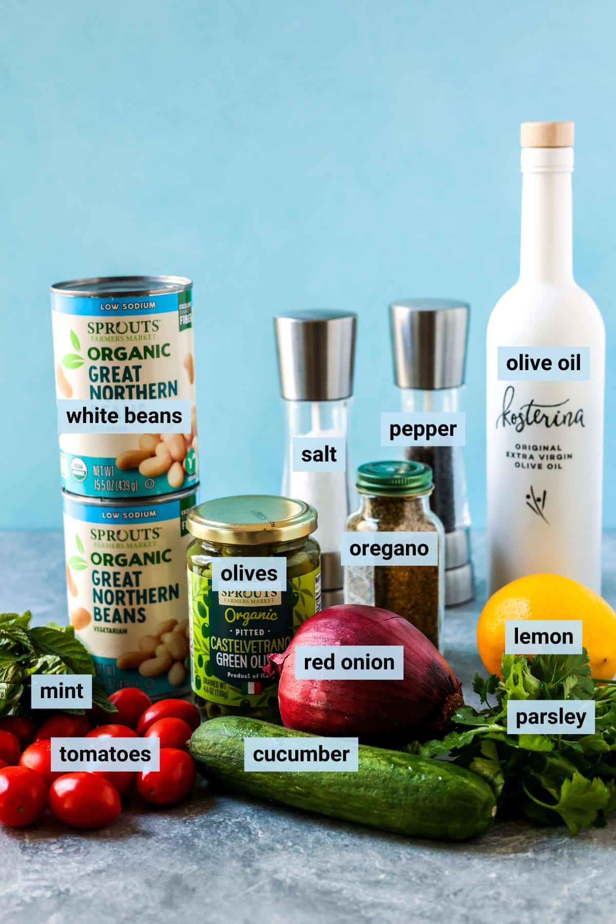Cans of great northern beans, fresh mint, grape tomatoes, cucumber, red onion, fresh parsley, a lemon, a jar of green olives, bottle of dried oregano, salt and pepper grinders, and a bottle of olive oil.