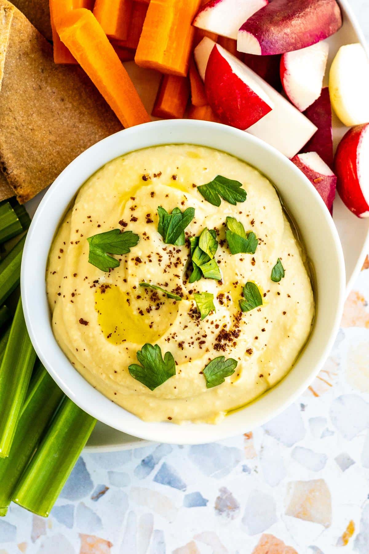 Bowl of hummus on a platter with pita bread, celery, carrots, and radishes.