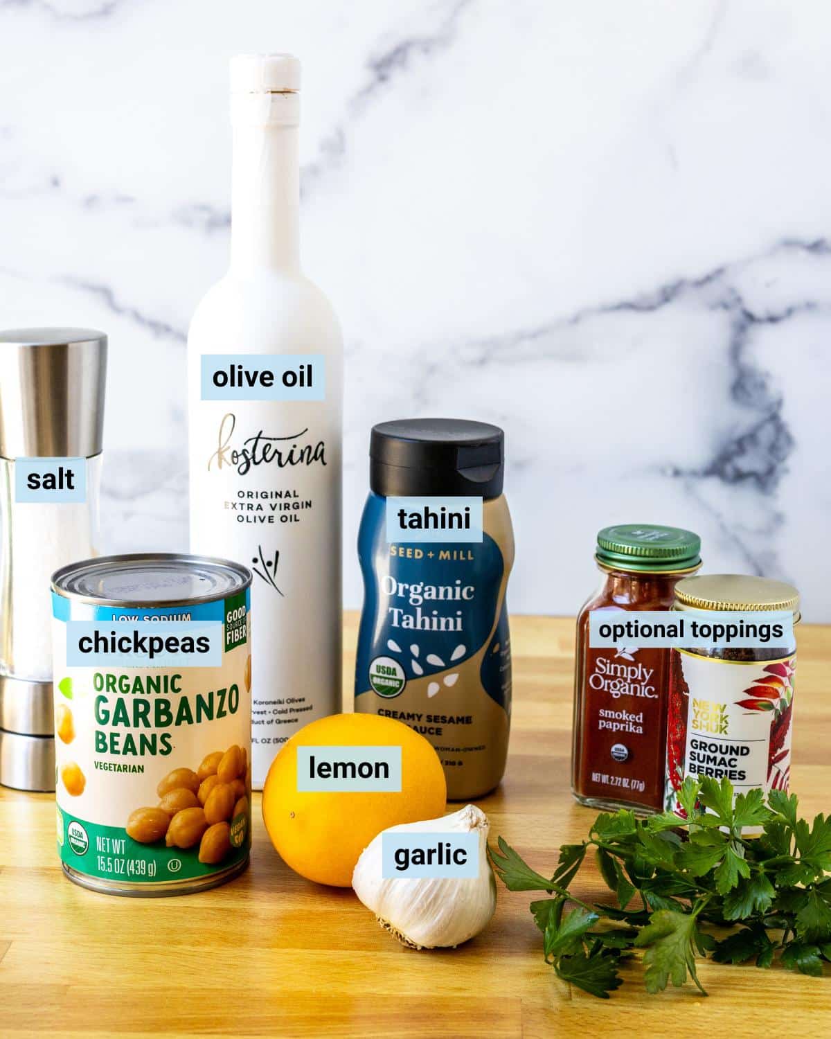 Can of chickpeas, salt grinder, bottle of olive oil, bottle of tahini, lemon, garlic, and optional toppings (paprika, sumac, and fresh parsley).
