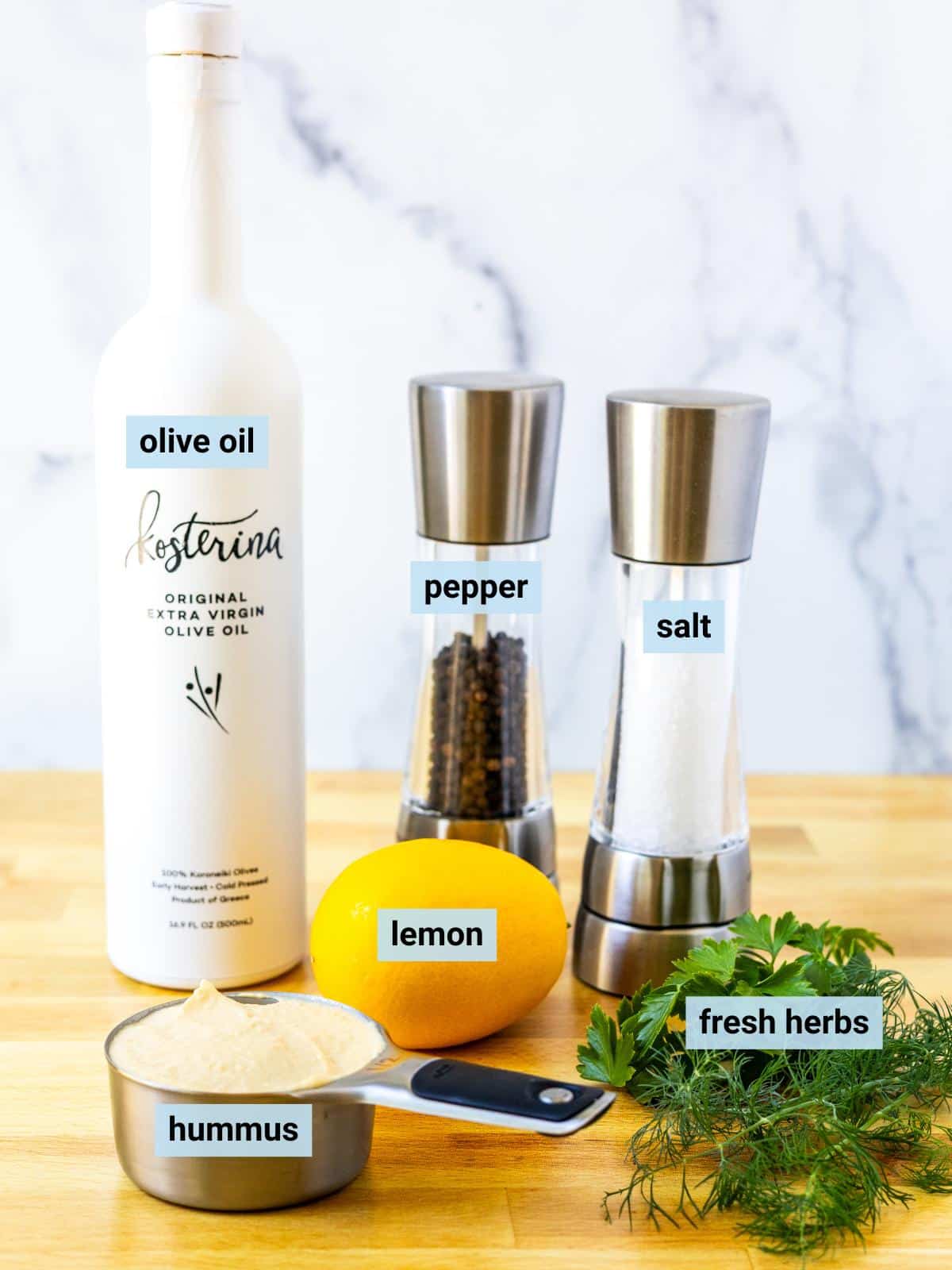 A measuring cup with hummus in it, a lemon, some fresh parsley and dill, a bottle of olive oil, and salt and pepper grinders.