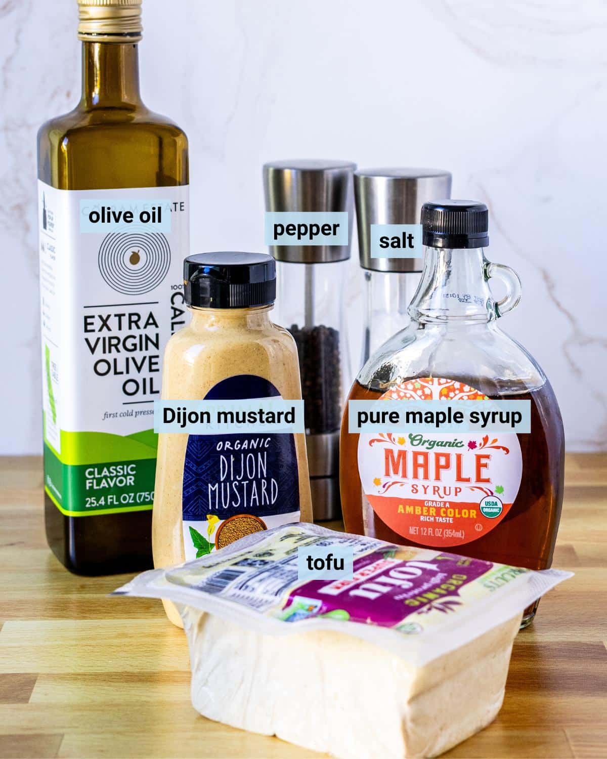 Block of super firm tofu, bottles of olive oil, Dijon mustard, and pure maple syrup, and salt and pepper grinders on a cutting board.