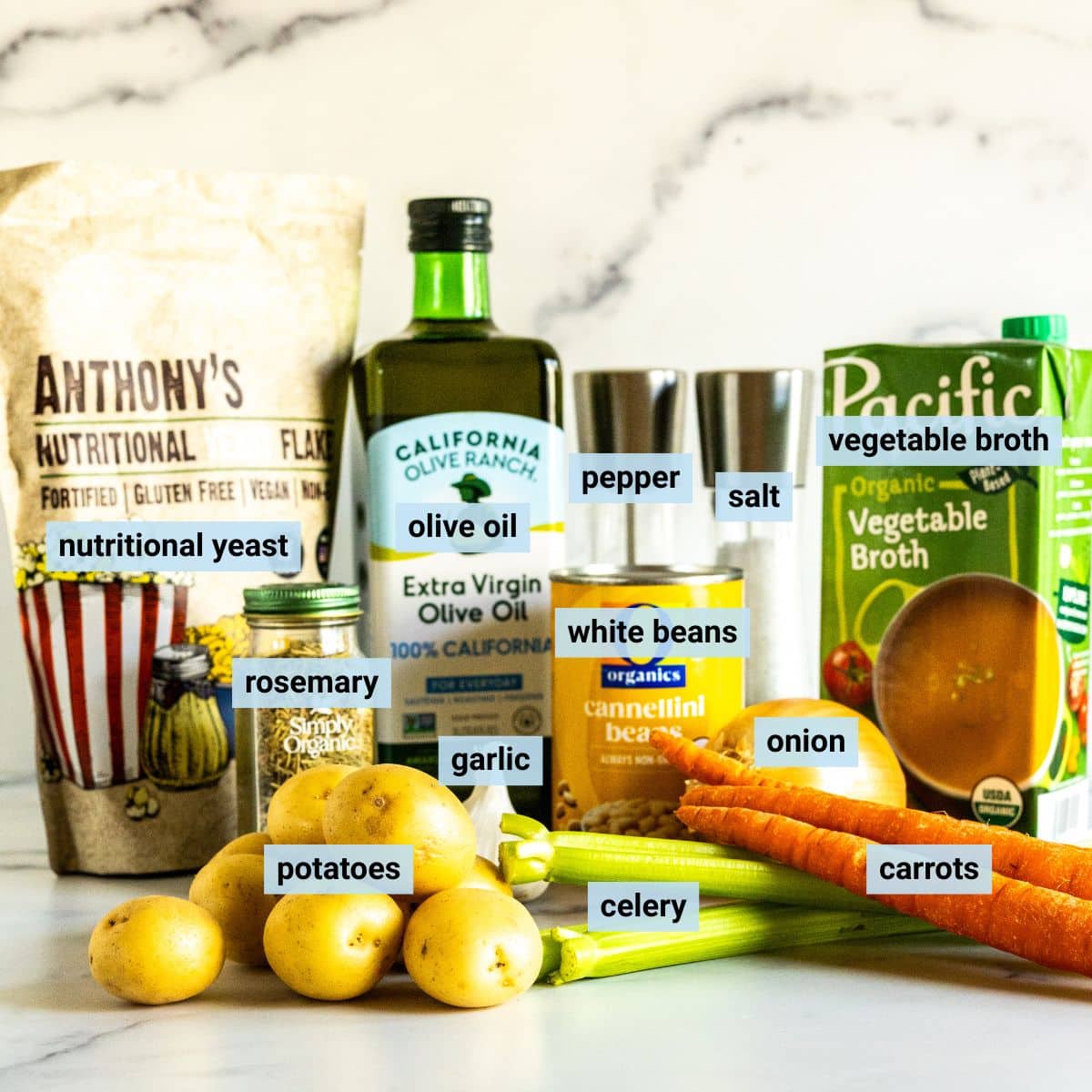 Labeled ingredients on a kitchen counter: yellow potatoes, celery, garlic, carrots, onion, jar of dried rosemary, bottle of olive oil, can of cannellini beans, box of vegetable broth, bag of nutritional yeast, bottle of olive oil, and salt and pepper grinders.