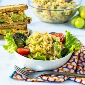 A green salad in a bowl with tomatoes and curried chickpea salad, a curried chickpea salad sandwich, and a bowl of chickpea salad and a bunch of green grapes on the side.