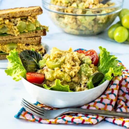 A green salad in a bowl with tomatoes and curried chickpea salad, a curried chickpea salad sandwich, and a bowl of chickpea salad and a bunch of green grapes on the side.