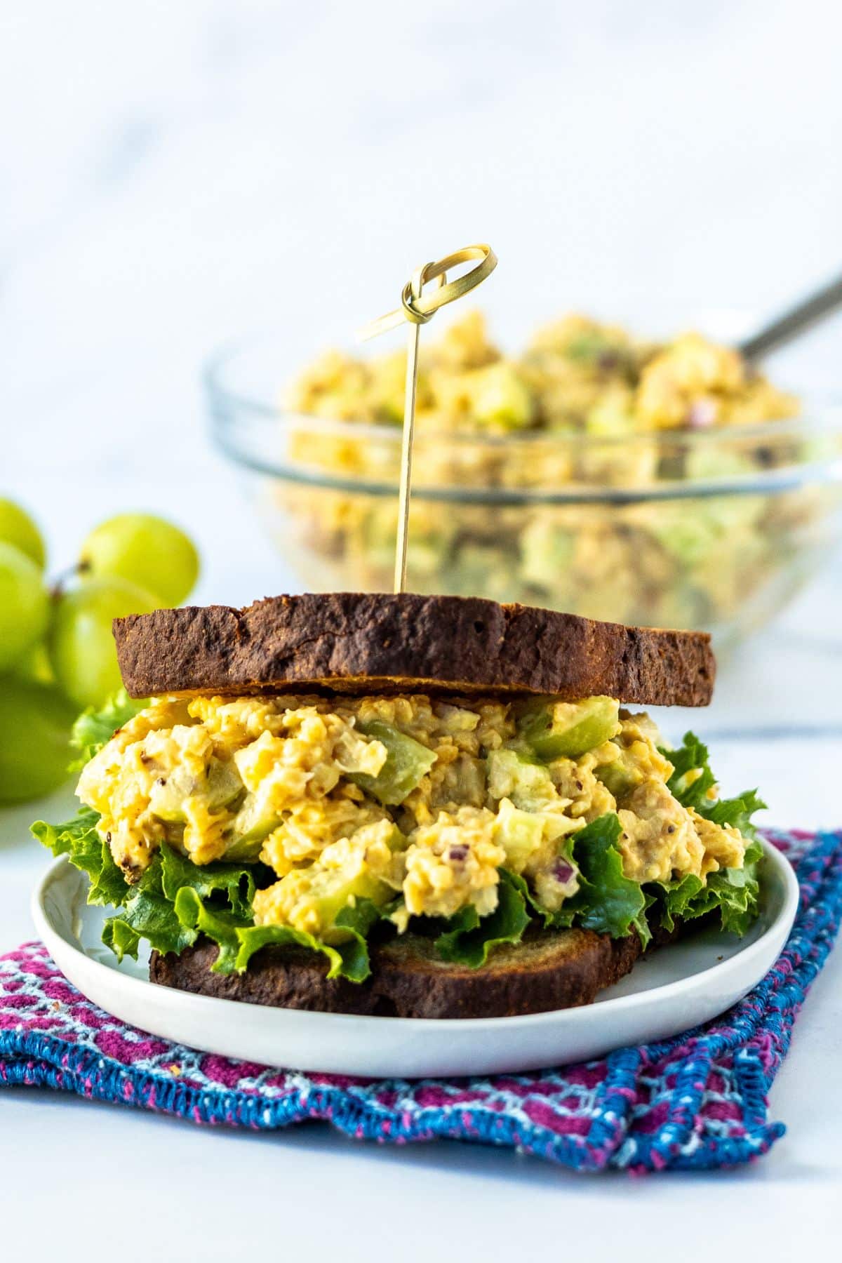 A chickpea salad sandwich on a plate with a bowl of the salad in the background and a bunch of green grapes.