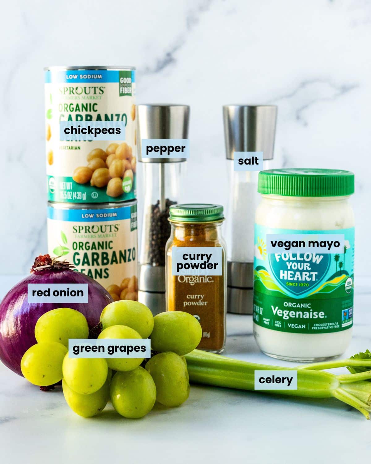 Cans of chickpeas, a red onion, a bunch of green grapes, celery ribs, a jar of vegan mayo, a bottle of curry powder, and salt and pepper grinders.